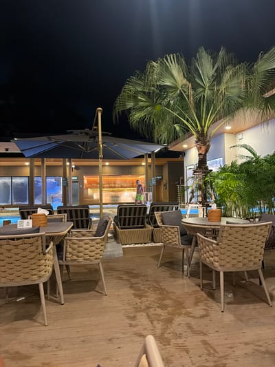 This image captures an outdoor seating area at night, likely a restaurant or bar, with a focus on the ambiance created by ambient lighting and tropical vegetation. The scene is set in Si Sunthon, Thailand, as indicated by the location context.
The primary subjects include several empty chairs and tables arranged on a wooden deck. The chairs are made of a woven material with a light beige color, and some have dark cushions. Palm trees are prominent, with one large palm tree dominating the left side of the frame and another cluster of green foliage to the right, suggesting a tropical setting. In the background, a building with large windows and warm interior lighting is visible. Through the windows, one can glimpse what appears to be an indoor swimming pool area with underwater lighting. A single person, a man wearing green swim trunks, is briefly visible standing near the pool.
The time of day is clearly night, indicated by the dark sky and the artificial lighting illuminating the scene. The wooden deck appears damp, suggesting recent rain or the presence of a pool nearby. The overall mood is relaxed and inviting, with the soft lighting creating a warm glow.
No specific activity or event is taking place; the setting appears to be after closing or during a quiet period.
Notable details include the intricate weave pattern of the chairs, the textured wooden deck, and the contrast between the dark night sky and the illuminated interior of the building. A sign on one of the tables reads "40 TABLE" along with a QR code, possibly indicating a table number or a menu access point. The wetness on the deck is a noticeable detail that adds to the atmosphere.