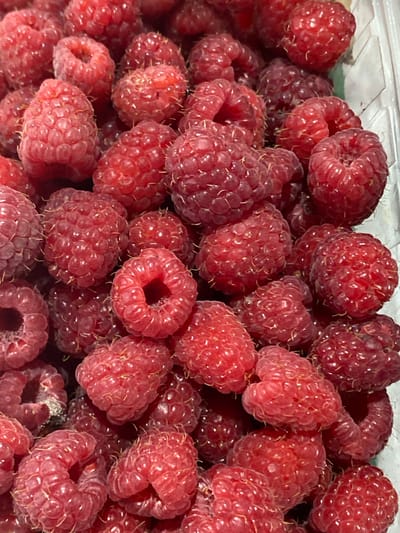 The image shows a close-up, top-down view of a pile of fresh, ripe raspberries. The berries are a vibrant red-pink color and have a textured, segmented surface. Some raspberries are whole, while others appear to be slightly compressed or misshapen. Fine, light-colored hairs are visible on the surface of many of the berries. The raspberries are densely packed together, filling the frame. The context of the city of Kushchovskaya, Russia is not discernible from the visual information present in the image. There are no people, actions, or environmental details beyond the berries themselves.