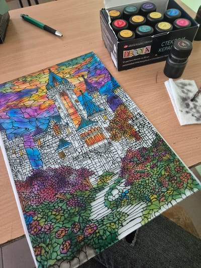 The image presents a crafting scene on a light brown wooden table. A partially opened box of "DECOLA" acrylic paints sits on the left, next to a black jar. Above the box lies a small, soiled white cloth and a green and black pen.
Occupying the majority of the table is a painting of a fantastical castle scene, reminiscent of stained glass. The castle has multiple towers, arches, and a complex network of white lines dividing the picture into small segments, filled with a variety of colors, including blues, purples, yellows, oranges, greens, and reds. The surrounding area features a dense, colorful garden or forest with vibrant flowers.
The image does not appear to be taken in Pavlikeni, Bulgaria.