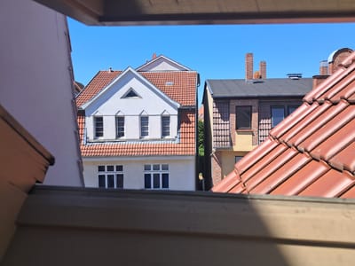 This image captures a view of rooftops and buildings in Parchim, Germany, seen through a framed opening that appears to be a window or attic space. The foreground features the edge of a wooden structure, likely part of the framing for the view. In the mid-ground, a white building with a red tile roof is prominent, featuring a triangular gable and several multi-paned windows. Adjacent to it, a narrower building with a dark grey roof and brick accents is visible. The background is dominated by a clear blue sky. The red tile roofs are a common architectural feature in the region. The scene appears to be an outdoor view on a sunny day, possibly from an upper floor or vantage point within a residential or urban area of Parchim. There are no people or animals visible in the frame, and the scene is static and observational. The overall impression is one of a quiet, residential street or neighborhood.