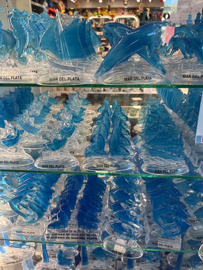 The image displays multiple clear glass shelves inside a souvenir shop located in Mar del Plata, Argentina. The shelves are densely packed with numerous translucent plastic figurines, primarily in shades of blue and clear/frosted white. These figurines are molded into various maritime shapes, including seahorses, sailboats, dolphins in jumping and intertwined poses, and sea lions. Each figurine is affixed to a clear, oval-shaped base, which uniformly bears the molded or printed text "MAR DEL PLATA"; some bases additionally display "RECUERDO DE MAR DEL PLATA" (Souvenir of Mar del Plata). Three levels of these displays are visible, with the background out of focus, indicating other retail merchandise in an indoor commercial setting. No individuals or active interactions are present in the scene.