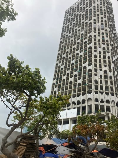 The image presents an urban landscape in Hải Vân Ward, Vietnam, under a uniformly overcast sky. A tall, slender concrete high-rise building, seemingly incomplete or under construction, occupies the majority of the right side of the frame. Its facade is characterized by a repetitive grid of numerous elongated, arch-shaped openings, which serve as window frames, revealing interior structural elements. Sections of green safety netting are visible within some of these openings. A white banner with "TELIA" visible in black text is affixed to a lower section of the building's exterior. The foreground and midground on the left feature multiple trees with green foliage. Below these trees, an area contains stacks of wooden pallets and various items covered by blue and orange tarpaulins, suggesting a makeshift storage or work zone. No individuals, explicit actions, or interactions are discernible within the scene.