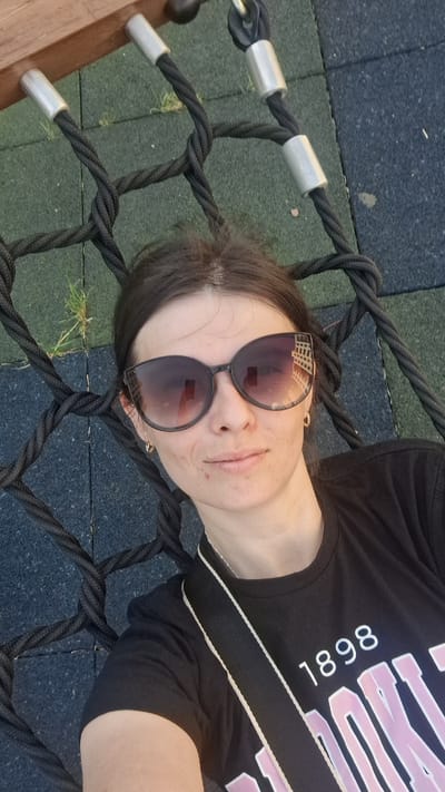 A woman is captured in a selfie, lying on a rope net structure, likely a swing or climbing net, on a playground. She is wearing oversized, dark-lensed sunglasses and a black t-shirt with white text that reads "1898" and some other pink lettering that is partially obscured. Her hair is dark and pulled back. The background consists of a black rope net and a playground surface made of interlocking rubber tiles in shades of dark blue and green. Some sparse blades of grass are visible between the tiles. The scene appears to be outdoors during daylight.