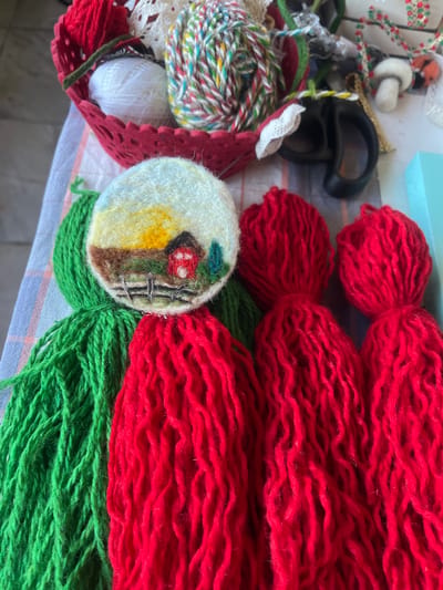 The image shows a close-up of various crafting materials, predominantly yarn and a small embroidered scene, laid out on a striped fabric surface. In the upper left, a red basket holds balls of white and multi-colored yarn, along with some white lace. To the right of the basket, a pair of black scissors is visible amongst other decorative items, including small felted figures. The central focus is a round, embroidered piece depicting a rustic landscape with a red barn, a fence, trees, and a setting sun with a yellow and orange sky. Below this embroidered piece are large bundles of bright green and red yarn, resembling tassels. The image is taken from a high-angle perspective, with shallow depth of field blurring the background elements. There is no discernible information about the city of Cheshnegirovo, Bulgaria, or any people present in the scene.