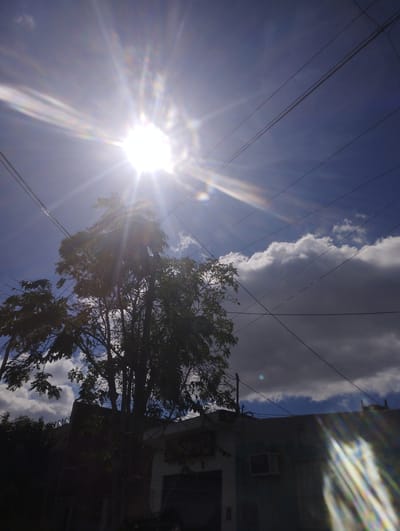 The image shows a daytime scene in Gregorio de Laferrere, Argentina. A bright sun dominates the upper left corner, casting intense rays across the sky. The sky is a mix of blue and clouds, with some clouds appearing dark and heavy. A tree is located in the center of the image, partially obscuring the sun. Electrical wires run across the sky, intersecting the tree and the sun's rays. In the lower portion of the image, a building is visible, partially obscured by shadows. An air conditioning unit is visible on the building. The scene is generally overexposed due to the bright sunlight.