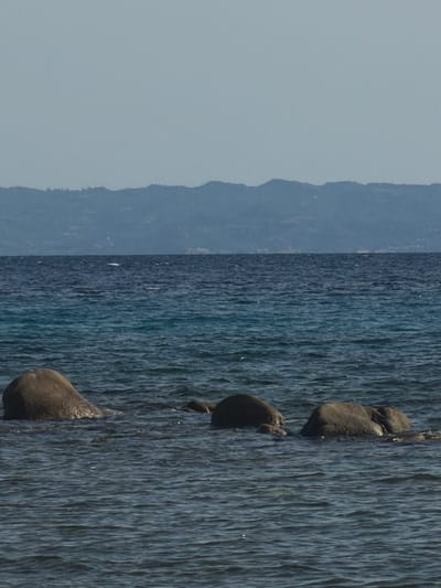 Three large, rounded rocks protrude from the calm, dark blue water of the Aegean Sea, near Sithonia, Greece. The rocks are partially submerged, with the surfaces that are exposed glistening in the sunlight. In the distance, a hazy, light blue mountain range forms the horizon line. The sky above is a pale, cloudless blue. The water exhibits subtle ripples, reflecting the clear sky.