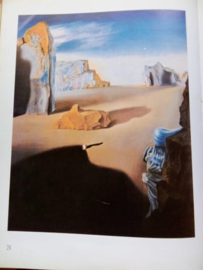 This is a painting of a surreal desert landscape under a cloudy blue sky. The ground is sandy and dry, with several large rock formations scattered throughout. In the foreground, a dark shadow covers a significant portion of the scene. A single, thin white object, possibly a stick or a staff, lies on the ground within the shadow. To the right, a dark rock formation juts into the frame, and partially obscured by it is a figure draped in flowing blue fabric. The figure appears to have a head with indistinct features and is seated or partially submerged in the rock. The overall impression is one of vastness, emptiness, and mystery, characteristic of Surrealist art. The image does not contain any elements that suggest a location in Zhmerynka, Ukraine.