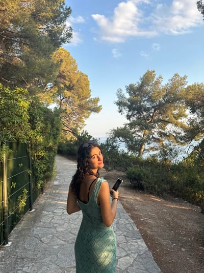 A woman in a light green dress stands on a stone path, holding a phone. Behind her is a wrought-iron fence with greenery beyond. The path leads through a wooded area with tall trees on either side. In the distance, a glimpse of the ocean is visible between the trees. The sky is blue with scattered white clouds. Sunglasses rest atop the woman's head.