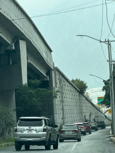 The image displays a traffic scene on an overcast day in Santa Catarina, Nuevo León, Mexico, specifically near Blvd. Diaz Ordaz. The perspective is from within a vehicle, looking ahead at a queue of cars on an asphalt road that appears damp.
In the immediate foreground, the rear of a silver Toyota 4Runner SUV (license plate PGP-2117) is visible. Following it is a grey sedan, then a line of vehicles including a red sedan, a dark SUV or truck, and a white commercial truck carrying ladders.
To the left, a large concrete elevated highway structure, supported by multiple robust concrete pillars, spans the upper portion of the frame. Green trees and bushes grow beneath and around this infrastructure. To the right of the road, a tall, textured retaining wall made of rectangular concrete segments runs parallel to the traffic. Further right, utility poles with overhead wires and streetlights are visible. A green road sign, partially obscured, displays "TRANSPORTES" and "Blvd. Diaz Ordaz," with a circular white speed limit sign below indicating "60" km/h. In the background, hazy mountains are visible beneath a uniform light grey sky, with what appears to be a residential area on a distant hillside beyond the retaining wall. No individuals are visible.