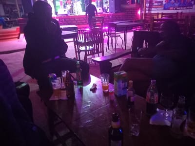 The image shows a dimly lit indoor space with a group of people seated at a table. The table is covered in various drinks and snacks. The people are seated in a relaxed posture, seemingly engaging in conversation. The environment is dimly lit, with only a few lights illuminating the scene. The furniture consists of wooden tables and chairs. The space is sparsely furnished with minimal decor. There are two men seated at the table, one with a dark jacket on and the other with blue shorts on. The table also contains four bottles of drinks, five glasses, and a few snacks. The scene suggests an informal gathering of friends enjoying drinks and snacks in the evening. The location is a bar or restaurant in Bungoma, Kenya, at the coordinates (0.593, 34.559).