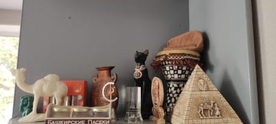 The image shows a shelf displaying various decorative items, including Egyptian-themed figurines and pottery, arranged in a room in Bogdashkino, Russia. In the foreground, a cream-colored camel figurine stands next to a small green bust. Behind them, an orange box and several jars are visible. To the right, a large, ornate brown vase with handles is positioned next to a black cat statue wearing a jeweled collar. A glass container with a depiction of a building is to the left of a drum-like object with a checkered pattern and fringe, topped with a knitted cap. A detailed pyramid figurine with hieroglyphics and carved figures is at the far right. The background consists of a plain gray wall, with a window and a textured wall visible on the left and right edges, respectively. No people are present in the scene.