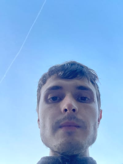 A low-angle, close-up, eye-level selfie of a young Caucasian male with brown hair and a mustache against a clear blue sky with a faint contrail. The man's face is the primary subject, positioned in the lower half of the frame. He is looking directly into the camera. The background consists entirely of a bright blue sky. No specific location in Genk, Belgium is discernible from the image.