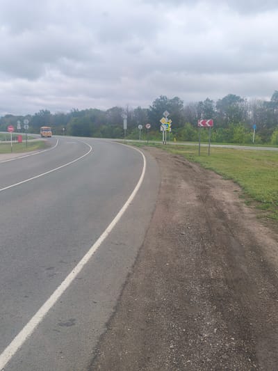 This image depicts a rural road scene in Zavolzh'e, Russia, under an overcast sky. The road curves to the right, with a white line marking the lane. To the left of the paved road is a dirt shoulder with tire tracks. A yellow bus is visible in the distance, driving on the road.
Several road signs are present. On the left, a red circular sign with a diagonal bar signifies "no entry." Adjacent to it are two other signs, one a diamond shape with a cross, and another a white rectangular sign with a blue figure. Further down the road, a red and white chevron sign indicates a sharp curve to the right. Another sign, a blue circle with a white arrow, is visible on the right side of the road.
The overall scene suggests a cloudy day, possibly late afternoon given the diffused light. The vegetation along the roadside is lush and green, indicating it's likely late spring or summer. The presence of the bus and road signs points to this being a publicly accessible road, likely in a rural or semi-rural area. There is no visible text on the bus or other discernible objects, except for the symbols on the road signs. The scene is calm and unremarkable, a typical view of a road in the Russian countryside.
