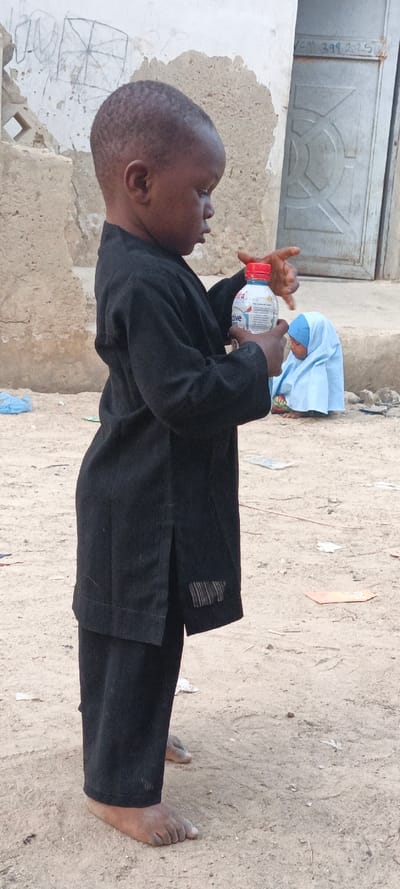 A young child, likely male, dressed in a black long-sleeved tunic and trousers, stands barefoot on sandy, light-brown ground in Rangaza, Nigeria. The child is captured in profile, facing towards the right side of the frame, intently looking down at a plastic water bottle held in both hands. The bottle has a red screw cap and a label prominently featuring the word "active" along with a colorful bird graphic.
In the mid-ground, a smaller child, partially obscured, is seated on the ground, wearing a light blue head covering. The background consists of weathered, light-colored walls; one wall behind the main child's head shows a faint, rough drawing resembling a square with diagonal lines. Further back and to the right, a grey metal door with an embossed circular pattern is visible, bearing faded text that appears to read "VCN 399 2025" near the top right corner. The ground is uneven dirt or sand, scattered with small pieces of debris. The scene is illuminated by natural daylight, indicating it is daytime. The overall setting suggests a humble, possibly rural or semi-urban environment.