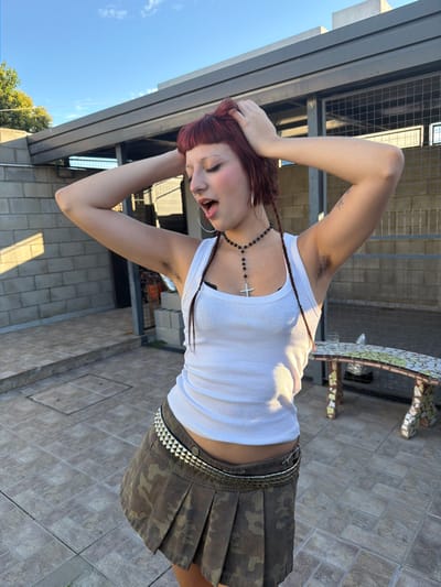 An individual is depicted standing outdoors on a tiled patio in Quilmes Oeste, Argentina, under a clear blue sky. The person, with reddish-brown hair styled with bangs and braids, wears a white ribbed tank top, an olive green camouflage-patterned pleated mini skirt, and a studded belt. A black beaded rosary-style necklace with a silver-colored cross pendant, large silver-colored hoop earrings, and a nose ring are visible. Both arms are raised with hands placed behind the head, revealing visible underarm hair. The individual's mouth is open, and eyes are partially closed. The background includes a light gray concrete block wall on the left, and a modern structure with a dark gray flat roof and metal framing on the right. A mosaic-tiled bench or table is partially visible behind the individual to the right. The scene is illuminated by natural light, casting shadows.