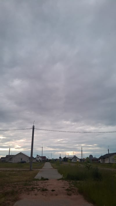 A vertical, eye-level, full shot captures a street scene in Stowbtsy, Belarus, under a heavily overcast sky. The sky dominates the upper two-thirds of the image, filled with dense, grey clouds that obscure the sun, though a faint luminescence suggests the horizon is near. A single, weathered utility pole with two horizontal wires stretches across the middle ground.
Below the wires, a paved sidewalk or path leads into the distance, flanked by a grassy verge on the left and overgrown weeds on the right. A few scattered houses, characterized by their pitched roofs and simple architecture, are visible on either side of the street. The houses appear to be single or two-story structures, with some exhibiting windows. A dark-colored car is parked in front of a house on the left. Streetlights are sporadically placed along the street. The foreground consists of unkempt ground with patches of dirt and dry grass. The overall impression is of a quiet, residential area on an overcast day, possibly at dusk or dawn.