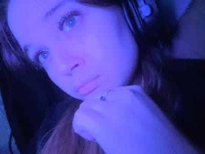 A young female is depicted in a close-up portrait, illuminated by a pervasive blue-purple light source. Her head is angled slightly to the right, with her blue eyes directed upwards and to the right. Her right hand is raised, with her chin resting on her knuckles. A silver-colored ring with a blue gemstone is present on her right ring finger. She wears dark over-ear headphones, with a light-colored ear cushion visible on the right side of her head. Her hair is dark brown or auburn. The background is dark and indistinct, suggesting an indoor environment. No specific location within Pushchino, Russia, is discernible from the visual information provided.