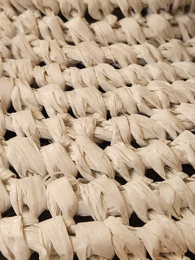 This is a close-up, angled shot of a woven material, likely a basket or a similar decorative item. The material appears to be made of thick, pale straw or raffia, tightly woven in a crisscross pattern. The lighting casts shadows that highlight the texture and depth of the weave, giving it a tactile quality. Some strands of the material show slight variations in color and texture, with minor specks of what could be dirt or natural imperfections in the straw. The overall impression is one of rustic craftsmanship. No people, activity, text, or specific location cues are visible beyond the general context of the United States of America, Maitland. The time of day and weather are not discernible from the image.