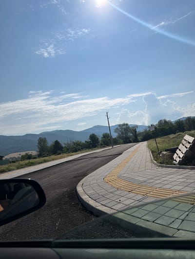 This is a photograph taken from inside a vehicle, looking out at a curving road under a bright, sunny sky with scattered clouds. The road is newly paved and bordered by a sidewalk made of light gray and yellow paving stones. In the background, rolling green hills and mountains are visible under the blue sky. A wooden pallet lies on its side on the grassy verge next to the sidewalk, and a utility pole stands near the road. The sun's rays create a lens flare effect in the upper right corner of the image. The scene appears to be located in a semi-rural area, possibly on the outskirts of the city of Pravets, Bulgaria.