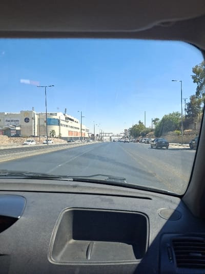 The image displays a forward-facing view from inside a vehicle on a multi-lane asphalt road in Amman, Jordan. The vehicle's dark grey dashboard, including a central storage compartment and a right-side air vent, occupies the lower foreground.
On the left side of the road, a large, light-colored commercial building complex is visible. This complex prominently features a yellow McDonald's "M" logo and "Max Minute" signage, identifying its location on Princess Basma Street, approaching the 7th Circle. Several white and light-colored cars are parked or situated near the building. Multiple black streetlights line this side of the road.
On the right side of the road, a vegetated hillside or embankment rises, containing green trees and bushes, with additional streetlights. The road itself shows moderate vehicle traffic, with cars present in both directions. In the mid-distance, a white pedestrian bridge spans the roadway. The sky is clear and blue, indicating a sunny daytime environment. No specific individuals are discernible in detail.