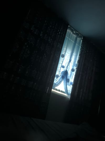 The image displays an indoor scene in Straldzha, Bulgaria, focusing on a window covered by multiple layers of drapery. The central element is a set of light-colored, likely sheer, curtains. These translucent curtains feature a distinct abstract pattern composed of interconnected light blue lines forming rectangular and circular shapes, some resembling spirals. The fabric is gathered and draped, creating soft folds that filter light from an unseen source beyond.
Above and below these sheer curtains are darker, opaque curtains or blinds, appearing black or very dark grey. These darker textiles also have a subtle, perhaps damask-like, pattern that is difficult to discern clearly due to the dim lighting in those sections. The overall lighting in the image is subdued and diffused, suggesting either an overcast day or early morning/late afternoon light filtering through the window.
The scene is static, with no human figures or discernible activity. The visible environment consists primarily of the window and its coverings, flanked by shadowed interior walls to the left and right. There is no visible text. The image highlights the interplay of light and shadow, and the decorative patterns of the window treatments.