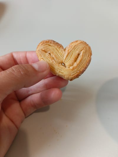 A hand holds a heart-shaped palmier cookie, dusted with granulated sugar. The cookie has a flaky, layered texture and a light golden-brown color. The background is a plain, light-colored surface. There are no identifiable landmarks or elements that would indicate a location in Malaya Vishera, Russia.