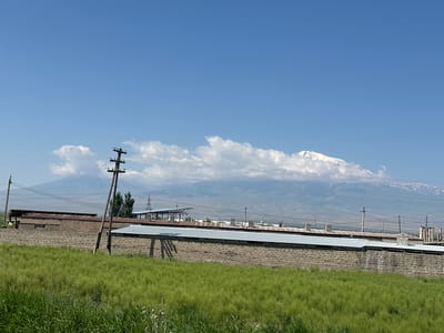 The image captures a daytime view of a rural landscape with industrial structures in the foreground and a majestic snow-capped mountain range in the background. The scene is set in the city of Shahumian, Armenia.

In the foreground, a dense field of vibrant green grass stretches across the lower portion of the frame. Behind the grass, a long, low building constructed from light-colored brick is visible. This building has multiple sections with corrugated metal roofs, some appearing weathered and rusted. A single, tall wooden utility pole with insulators stands prominently to the left of the building, with power lines stretching across the scene. Another utility pole is visible further to the left, and a few more industrial structures with similar roofing are scattered throughout the mid-ground.

Dominating the background is a vast mountain range, partially obscured by a band of white clouds. The peaks of the mountains are covered in snow, creating a stark contrast against the clear blue sky. The overall impression is one of a rural setting with agricultural land and industrial elements juxtaposed with the grandeur of the natural landscape. The sky is clear and blue with a few scattered white clouds.
