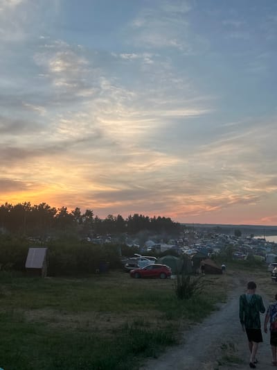 The image depicts a large outdoor gathering, possibly a festival or camping event, at dusk in the vicinity of Жемчужный, Russia. In the foreground, two young males, dressed casually, walk away from the camera on a dirt path. The middle ground features a campsite with numerous tents and cars, situated behind a line of trees. The sky above is filled with dramatic clouds, illuminated by the setting sun, casting orange and pink hues. The overall scene suggests a lively event taking place in a natural setting.