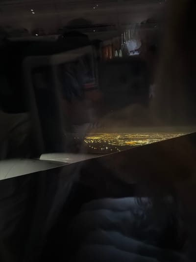 The image shows a night view from an airplane window, looking out at a sprawling cityscape illuminated by countless lights. The city appears to be situated along a coastline, with water visible in the distance. The lights are a mix of yellow and white, indicating a densely populated urban area. The airplane wing is visible in the foreground, partially obscuring the view. The reflections in the window suggest the interior of the aircraft, with faint outlines of passengers and cabin lights. The overall impression is of a vast and vibrant city seen from above at night.
The description does not contain enough specific detail to identify the location as Üsküdar, Turkey.