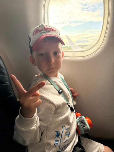 A young boy sits by an airplane window, wearing a white and red baseball cap, a grey sweatshirt with an anime-like illustration and Japanese text, and a light blue lanyard with a small orange and grey camera attached. He is holding up two fingers in a peace sign. Outside the window, the sky is filled with clouds and a portion of an airplane wing is visible. The boy is positioned in the foreground, with the airplane cabin and window filling the background. There is no information about Murmashi, Russia in this image.