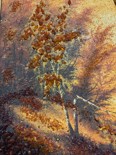This is a close-up, detailed photograph of a textured artwork. The artwork features a depiction of a tree with visible branches against a background that transitions from dark, textured earth tones to lighter, golden-yellow hues. The texture is enhanced by small, irregular pieces of amber or similar translucent resin scattered across the surface, particularly clustered around the tree and in the lower left portion of the image. These amber pieces are of varying sizes and shades of orange and brown, some appearing polished and others rougher. The background itself has a grainy, mottled appearance, with speckles of dark and light particles. The overall impression is one of an abstract or mixed-media representation of a natural scene, possibly a forest or tree in autumn. The location of Panovka, Russia, is not visually discernible within the image itself; this detail appears to be external information.