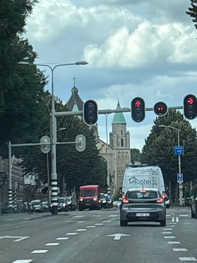 The image shows a street scene in Maastricht, Netherlands, with a focus on traffic and architecture. In the foreground, a grey car with license plate 2-GNZ-603 is driving on a multi-lane road. To the right of the car, a white van with the logo "Ropteco" is visible. A red truck is positioned further ahead in the left lane. Several other vehicles are present in the background, lined up at a traffic light.

Traffic signals are prominently displayed, with several showing red lights, indicating a stop. One signal displays a red arrow pointing to the left, another a red arrow pointing upwards, and another a red arrow pointing to the right. There are also pedestrian crossing signals visible.

In the background, a large, old stone building with a green-domed tower and a cross on top is the most prominent architectural feature, likely a church or cathedral. Trees line the street, and overhead utility poles and streetlights are visible. The sky is cloudy, suggesting overcast weather. The street itself is paved and marked with white lines.