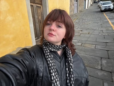 An individual with shoulder-length reddish-brown hair and bangs, wearing a black leather jacket and a black and white knit scarf, is depicted in the foreground, facing forward. Their left nostril exhibits a small piercing. The person's arm is partially visible on the bottom left, suggesting a self-portrait. To the immediate left of the individual is a textured, vibrant yellow stucco wall with areas of peeling paint. A weathered, dark brown wooden door with an ornate handle is visible behind the subject, set into a grey frame. To the right, a cobblestone street, composed of irregular grey flagstones, slopes upwards into the distance. A faded yellow line is painted along the right side of the street. Several light-colored parked vehicles, including at least two white SUVs, are visible further up the street on the right. Buildings with stone and stucco facades line the street. This scene is located in a narrow, uphill street within the city of Cortona, Italy.