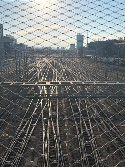 The image captures a wide view of a complex railway junction in Zurich, Switzerland, as seen through a wire mesh fence. The sun is setting, casting a warm glow across the sky and glinting off the metal rails below. The railway tracks form an intricate network, with numerous switches and crossings disappearing into the distance. In the background, the Zurich skyline is visible, featuring modern buildings and a distinctive tall tower. The foreground is dominated by the diamond pattern of the wire mesh, which creates a layered effect and partially obscures the scene. There are no people visible in the image. The overall impression is one of urban infrastructure and the vastness of the railway system.