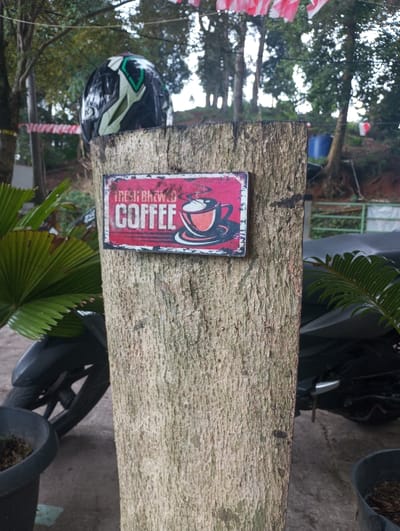The image displays an outdoor scene located in Gadog, Indonesia. A vertical, weathered wooden post stands prominently in the foreground. Affixed to the post is a rectangular, distressed red sign featuring white text that reads "FRESH BREWED COFFEE" and a graphic of a steaming coffee cup. Resting on top of the post is a black and green motorcycle helmet. To the immediate left of the post, a portion of a black motorcycle, including its front wheel and fender, is visible on a concrete or paved surface. Green foliage, including large palm-like leaves, is present in the lower left and right foreground. The background is dominated by numerous trees and undulating terrain. Red and white striped banners are strung horizontally between trees in the upper background, and a blue barrel is discernible further in the distance. No individuals are present, and no actions or interactions are depicted.
