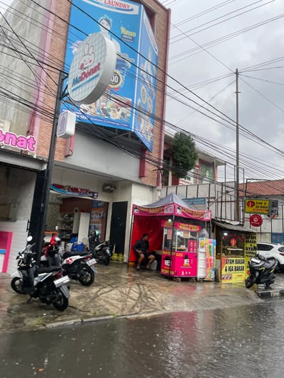 The image shows a street scene in Cimahi, Indonesia (latitude: -6.883, longitude: 107.552). The street is wet from recent rain, and there are several motorcycles parked on the side of the road. A man sits on a small stool in front of a food cart selling "Tahu Krispy" (crispy tofu), wearing blue shorts and a black shirt. The cart is pink and white with a black logo. Several other signs are visible, including one advertising a laundry service, another for a "McDonut" shop, and a third for "Ayam Bakar & Iga Bakar" (grilled chicken and ribs). The buildings in the background are brick with grey roofs.  There are power lines running along the street, and the sky is overcast with rain. There is a small, brightly colored sign for a "gofood" food delivery service.