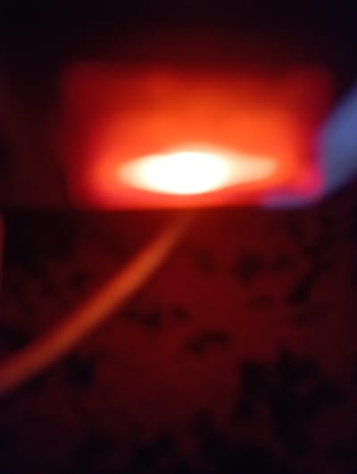 The image shows a close-up, out-of-focus view of a red light source, possibly an LED, emitting bright white light in the center. The light source appears to be housed within a red casing, with a slight blueish-white glow visible to the right. Below the light source, a blurry, elongated, light-colored object, possibly a cable or a pipe, runs diagonally across the frame. The background is a dark, reddish-brown color with indistinct, darker circular shapes, suggesting a textured surface like fabric or wallpaper. The overall impression is of a dimly lit environment with a dominant red and orange hue. The image is too abstract and lacks detail to determine its specific location within Ezbet Michel Hamawi, Egypt, or to identify any people, specific objects, or actions.