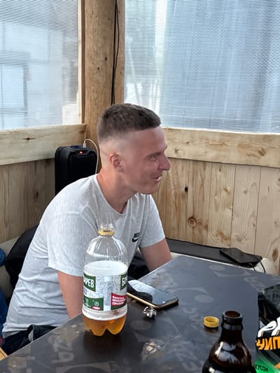 In an outdoor, covered seating area, a light-skinned male with a buzz cut and a grey t-shirt sits at a dark, patterned table. He is facing to his right, with a slight smile. On the table are a plastic bottle with a light brown liquid and a label in Russian, two cell phones, a small silver ring, a yellow bottle cap, and a dark brown glass bottle. Behind the man, a black portable speaker is visible. The background consists of wooden plank walls and a translucent, ribbed plastic wall. The location is identified as Бородулино, Russia.