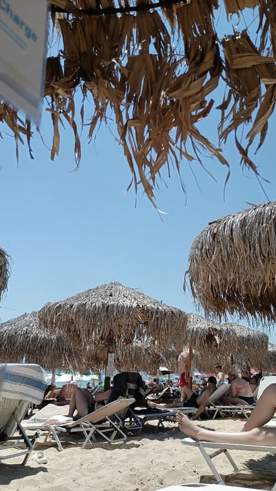 This image depicts a sunny day at a beach in Nea Peramos, Greece, under a clear blue sky. The foreground is dominated by sandy beach chairs and umbrellas with thatched roofs, providing shade. Several people are seen relaxing on the beach chairs, some are partially visible, others are fully reclined. In the background, more beach umbrellas and loungers are visible, along with other beachgoers and possibly some structures along the coastline. The overall scene conveys a typical beach resort atmosphere.