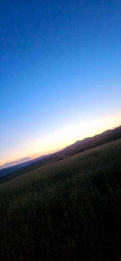The image shows a landscape scene at dusk, likely in the vicinity of Momin Prohod, Bulgaria. The sky transitions from a deep blue at the top to lighter shades of blue, pink, and orange near the horizon, indicating the sun has recently set or is about to rise. Rolling hills and mountains are visible in the distance, silhouetted against the lighter sky. In the foreground, a field of tall grass or crops is dark and textured, suggesting it is nighttime or late twilight. The overall impression is one of a tranquil, natural environment during the transitional hours of the day. There are no people or man-made structures clearly visible.