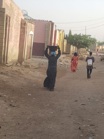 The image captures a dusty street scene in Dutsinma B, Nigeria. A woman wearing a long, black and white patterned dress and a blue headscarf is carrying a square, dark-colored object on her head. Further up the street, a woman in a red and orange patterned garment walks ahead of a person carrying a white cylindrical object on their shoulder. Buildings of varying heights and colors, primarily in earth tones, line the left side of the street. The street is unpaved and covered with dirt and debris. Overhead electrical wires stretch across the sky. The light is bright, suggesting a daytime scene.