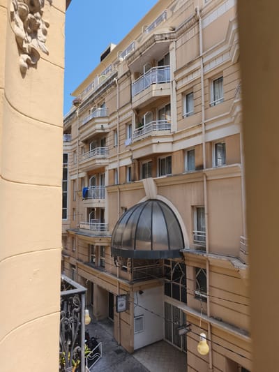 The image shows a section of a tan-colored building in Beausoleil, France, seen from an elevated vantage point. The building has multiple stories with balconies featuring glass railings and white trim. Some balconies have plants and laundry drying. The architecture includes decorative moldings and recessed window frames. A large, semi-circular glass canopy covers an entrance area on the ground floor, which has a patterned tiled floor and several doors. A black wrought-iron fence is visible in the foreground on the left, with a yellow decorative light bulb hanging from it. The sky is clear and blue. The image captures a street-level view of the building's facade and entrance, with a clear, sunny sky overhead.