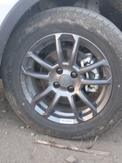The image shows a close-up of a car's wheel and tire. The wheel is dark gray with a multi-spoke design. The tire appears somewhat dirty and worn, with some dirt and debris visible on its surface. The brake caliper is visible behind the wheel spokes and is a light silver color. The surrounding environment appears to be unpaved ground, with some small rocks and a twig on the ground in the foreground. The image is taken from a slightly low angle, focusing on the wheel assembly. The location is not determinable from the image itself, and no information about Kandyagash, Kazakhstan is present.