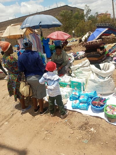 The image depicts an outdoor market scene in Tala ward, Kenya, under a bright, partially cloudy sky. In the foreground, a vendor, wearing an olive-green shirt and apron, crouches behind a display of merchandise laid out on a white tarp on the dusty ground. The stall features several large white sacks, numerous packaged diapers (brands "Mummy Baby" and "Muva," including "XL Large" sizes), and two small baskets containing various colorful small items, possibly hair accessories or trinkets.

Three individuals stand facing the vendor: two adult women and a young child. The woman on the far left wears a multi-colored patterned dress and an orange knitted hat, carrying a light-colored woven handbag. The woman next to her wears a blue patterned top, grey shorts, and a dark headscarf. A young child, wearing a red cap, a white and blue striped long-sleeved shirt, and green pants, stands directly in front of the vendor, facing the products.

Behind the immediate interaction, a blue SportPesa branded umbrella provides shade over a clothing rack with blue garments. A beige umbrella and a red umbrella are also visible, shading other market areas. The background shows an expansive open-air market with numerous goods, including stacks of fabric, mattresses, and other items spread on the ground. Several other marketgoers and vendors are present in the midground. Structures made of stone or brick are visible in the far background, along with some sparse vegetation. The ground throughout the market is dry, light-brown soil.