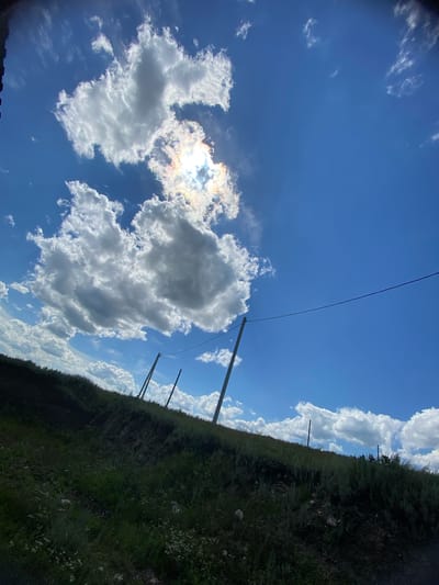 A low-angle, wide shot captures a bright, partly cloudy sky above a grassy hillside. The sun is visible, partially obscured by a large, fluffy cloud, casting a halo of light that refracts into subtle rainbow colors. A thin, dark wire stretches horizontally across the upper portion of the frame, supported by utility poles. The hillside is covered in dense, green vegetation, with some scattered white wildflowers. The overall scene suggests an outdoor, possibly rural setting in the city of Yuldybayevo, Russia.