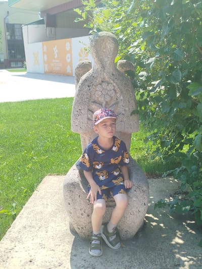The image shows a young boy sitting on a stone sculpture in Yalutorovsk, Russia. The boy is wearing a blue t-shirt with a tiger print and a colorful cap. He is sitting on a stone sculpture that appears to be a seated figure, with intricate carvings on its chest. The sculpture is located outdoors, with a grassy area to the left and lush green foliage to the right. In the background, there is a building with a white wall adorned with orange banners featuring various symbols and text that reads "Ялуторовская МАСТЕРСКАЯ СИБИРИ" (Yalutorovskaya MASTERSKAYA SIBIRI), indicating the location as the Yalutorovskaya Masterskaya Sibiri (Siberian Workshop of Yalutorovsk). The scene is illuminated by bright sunlight, casting shadows on the ground.