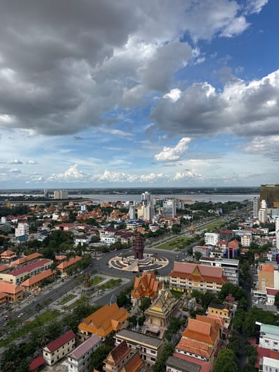 This is an aerial, rotated photograph of a cityscape in Khan Boeng Keng Kang, Cambodia. The image is divided diagonally, with the left side dominated by a cloudy sky, transitioning to a clear blue sky with scattered white clouds. The right side showcases a densely populated urban area situated along a wide river.
The city comprises a mix of modern buildings and traditional architecture, characterized by terracotta-tiled roofs. Prominent features include a circular plaza with a central monument, surrounded by trees and a multi-lane road. Numerous vehicles, including cars and motorcycles, are visible on the roads, indicating moderate daytime activity. Lush green trees are interspersed throughout the urban landscape, lining the streets and parks.
The time of day appears to be midday or early afternoon, given the bright lighting and the nature of the shadows. The weather is partly cloudy, with a mix of dark storm clouds and lighter, brighter clouds. The river is wide and appears to be a significant waterway, with a few boats visible on its surface. The overall scene depicts a bustling urban environment in a tropical or sub-tropical setting. No text is legible in the image.