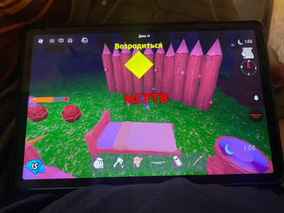 The image is a screenshot of a video game displayed on a tablet device. The game appears to be a survival or role-playing game with a cartoonish art style. The screen shows a colorful environment with a green grassy ground and a wooden fence in the background. In the foreground, there is a bed, two red mushroom-like objects, and various game interface elements.
Specifically, the game screen displays:
- At the top center, the text "День 97" (Day 97) and "Возродиться" (Respawn) in yellow text. Below this, a yellow diamond shape is prominent against the wooden fence.
- The text "МЕРТВ" (DEAD) is displayed in large red letters across the middle of the screen.
- On the left side, there's an orange health bar with a red figure at the end, and a blue diamond shape with the number "15" on it.
- A row of numbered item slots is at the bottom, showing icons for a bag, an axe, a pink object resembling a tool, and a bottle.
- On the right side, there is a compass and a clock showing "1:06" and a crescent moon icon.
- A red button with white text "Перестать бегать." (Stop running.) is in the upper right corner.
The tablet is placed on a surface that appears to be a dark fabric, likely a blanket or clothing, with some fuzzy texture. The lighting suggests the image was taken indoors in a dimly lit environment, with the tablet screen being the primary light source. There are no identifiable people or specific city locations visible in the image. The game's content does not provide any clues to its location within the city of ЖК «Борисоглебское», Russia, or any other specific geographic location.