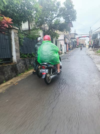 The image shows two people riding a motorcycle on a wet road in Cimenyan, Indonesia. The person driving the motorcycle is wearing a bright green raincoat and a red helmet. The other person riding on the back is wearing a dark jacket. Both people are not visible from the face due to the angle of the image. They are traveling along a narrow, paved road lined with buildings on either side. There are trees and vegetation in the background. The road is wet from recent rain, and the street is mostly empty. The motorcycle is moving forward at a moderate speed. The license plate of the motorcycle can be seen at the back of the image as DL 3040 BK.