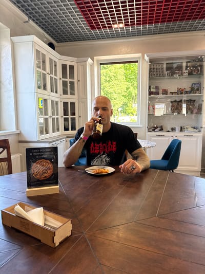 A bald man with tattoos on his arm sits at a large wooden table in a brightly lit cafe, holding a bottle of beer to his lips and drinking. He wears a black t-shirt with a red graphic design. On the table in front of him is a small plate with a slice of pizza and a fork. To his left, a stand displays an advertisement for "delicious and hot pizza from the oven" with an image of a pizza. A wooden napkin holder filled with napkins is also on the table. In the background, a blue chair is visible, and behind that, a glass-fronted cabinet displays various items, including Beatles memorabilia, figurines, and shoes. The cafe features white cabinets with glass doors along the wall and a large window offering a view of green trees. The ceiling has a grid pattern with red accents. The overall scene suggests a casual dining experience in a cafe located in the city of Radashkovichy, Belarus.